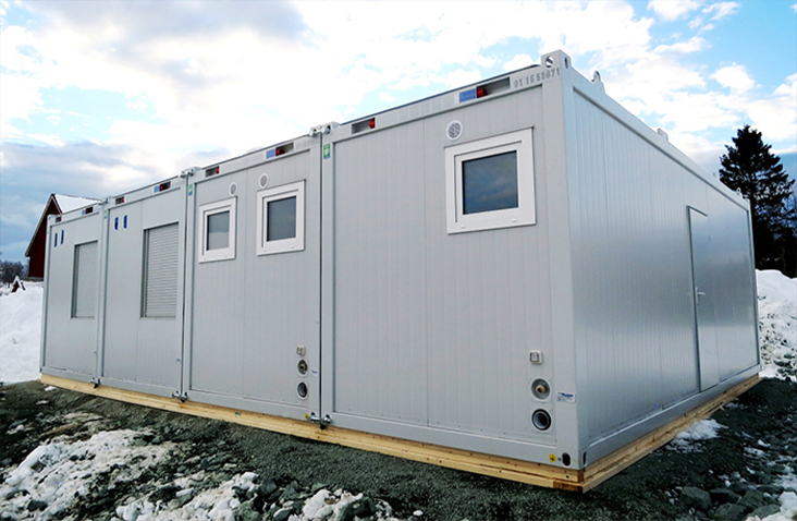 Large silver modular building made from four connected units with multiple windows and vents, installed on a raised timber base surrounded by snow
