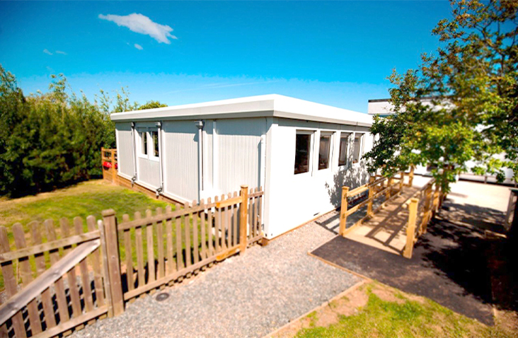 White modular school building with multiple windows, surrounded by fencing and trees, featuring an accessible ramp and gravel pathway under a bright blue sky.