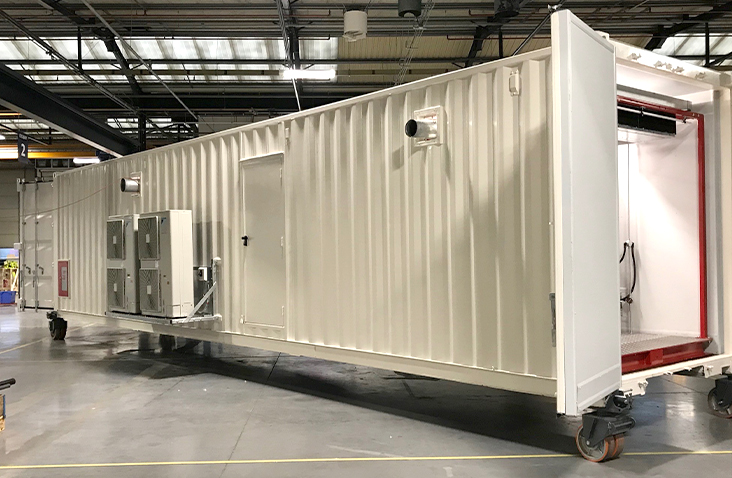 40ft white containerised equipment enclosure on wheeled supports inside a workshop. The unit features externally mounted HVAC systems, a personnel access door, and open cargo doors revealing internal plant space.