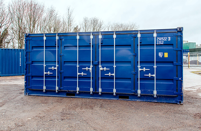 Blue 20ft side opening shipping container with closed side doors, positioned on a gravel surface in a storage yard with trees and fencing in the background