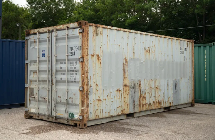 Weathered 20ft shipping container in pale grey, showing extensive rust along the edges and sides, with closed cargo doors, positioned on a yard surface with trees in the background. 