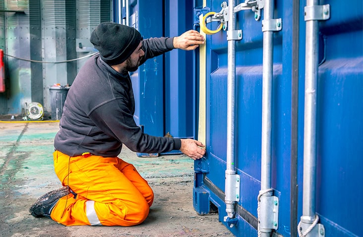 Engineer kneeling beside a blue shipping container and applying masking tape around the door edges before repainting. The worker wears orange high-visibility trousers, gloves, and a dark sweatshirt. 