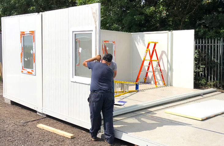 Workers assembling a flat pack office unit on site, showing panels with windows being installed during construction. 