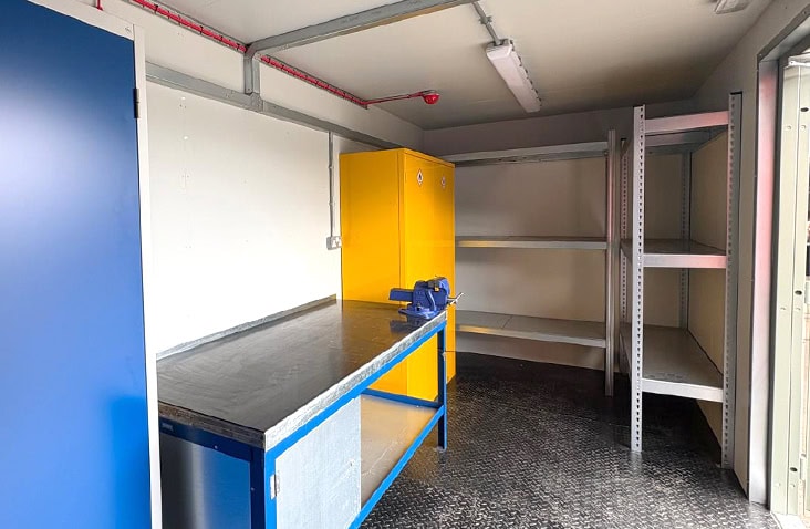 Interior of a converted workshop container with a metal workbench, shelving, blue and yellow tool cabinets, and fitted lighting. 