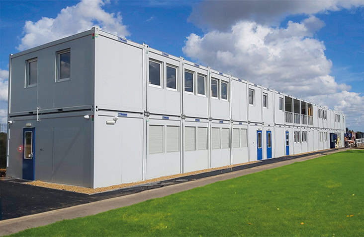 Two-storey modular school building made from stacked white units with blue doors and multiple windows, providing additional educational space beside a grassy area.