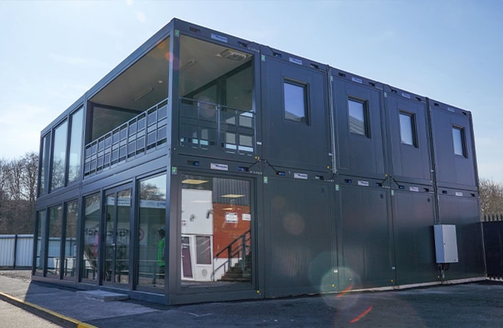 Modern two-storey modular office building made from stacked dark grey container units with large glass panels on the ground floor and smaller windows above.
