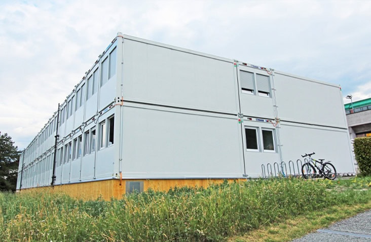 Large two-storey modular building made from multiple white units with windows, installed on a raised timber base beside a grassy area with bicycle stands.