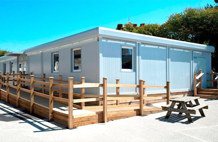 Single-storey modular school building with light grey panelled walls, multiple windows, and wooden accessibility ramps and fencing, located in a sunny schoolyard