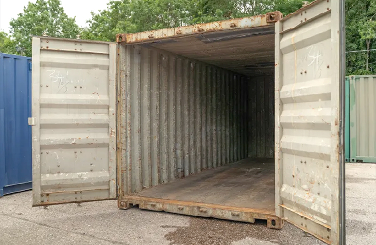 Weathered 20ft shipping container in pale grey with both cargo doors open, showing a worn steel interior and rust along the door frames and floor edges, positioned on a yard surface with trees in the background. 
