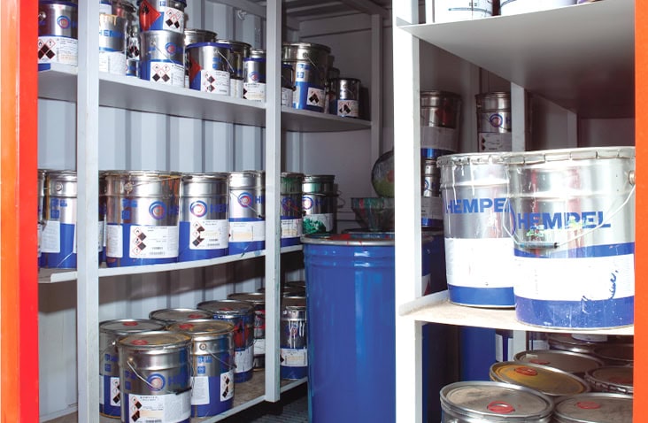 Shelving inside a shipping container storing numerous metal paint tins and a large blue drum, arranged for controlled chemical storage. 