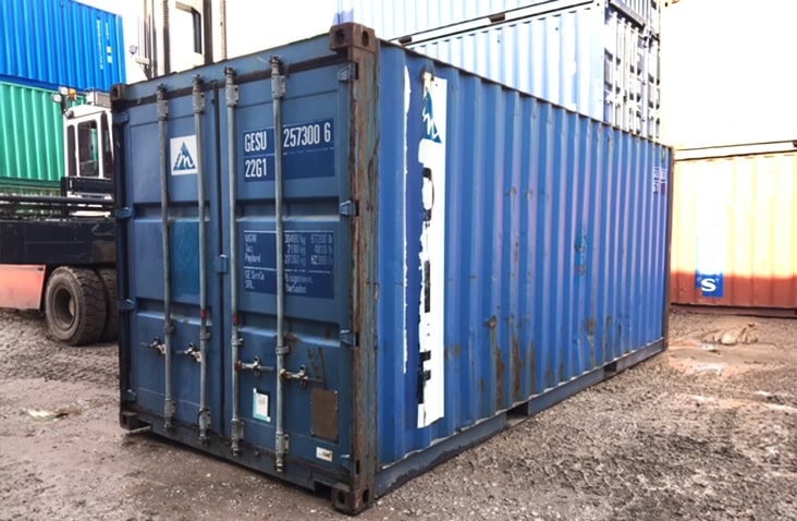 Used 20ft blue shipping container with closed cargo doors, showing areas of rust and worn paint, positioned on a rough yard surface with other containers in the background. 