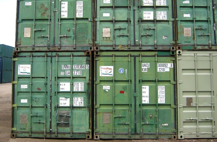 Stack of used green shipping containers with visible scratches, rust patches, and shipping labels, arranged in two rows in a container storage yard. 