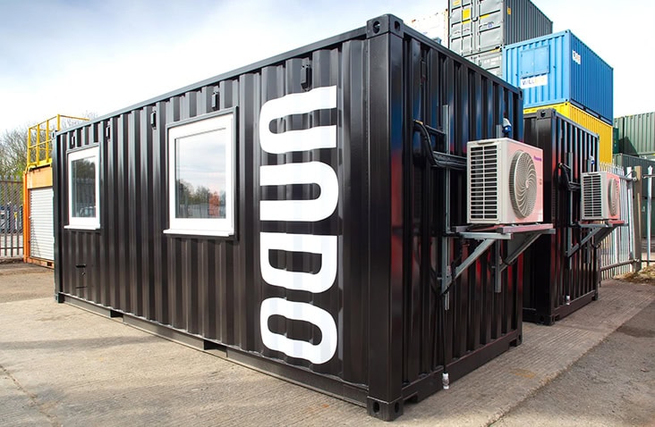 Black steel shipping container with large white ‘UNDO’ branding on the side, featuring two windows and external air conditioning units mounted on brackets. The container is placed outdoors on a concrete surface, with other stacked containers visible in the background under a clear sky.