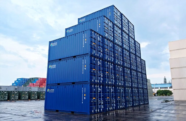 Stack of blue new 20ft shipping containers branded with S Jones Containers, arranged in a stepped formation at an outdoor container depot under a cloudy sky. 