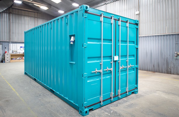 Bright turquoise steel shipping container with corrugated sides and closed double doors featuring silver locking bars. The container is placed indoors on a concrete floor within a large industrial workshop with corrugated metal walls and overhead lighting.