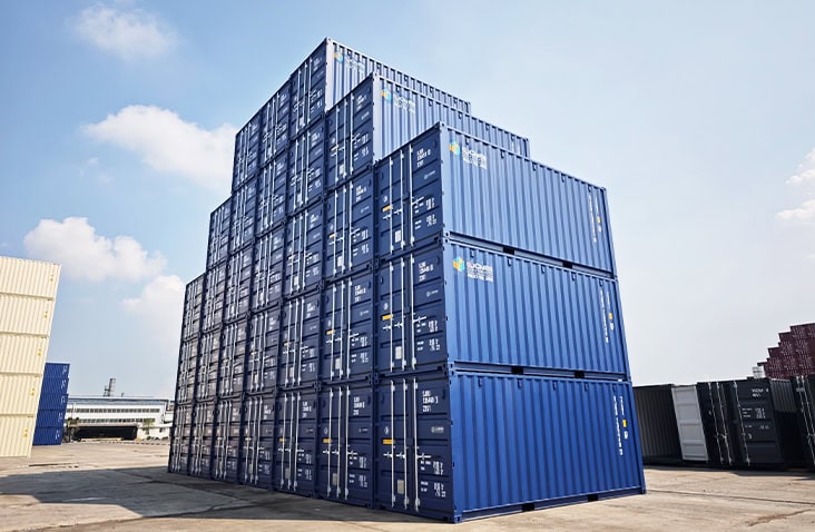Large stack of blue 20ft shipping containers arranged in a stepped formation at an outdoor storage depot under a bright sky.