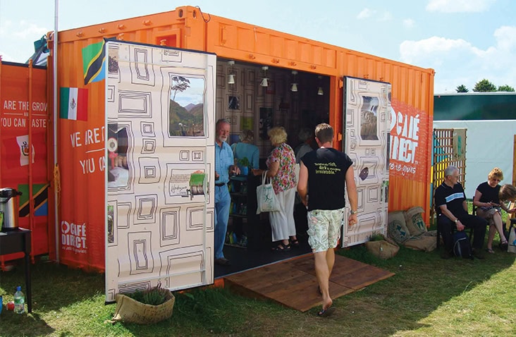 Bright orange shipping container converted into a café with open double doors featuring decorative panels, set up outdoors at an event with people entering and sitting nearby.