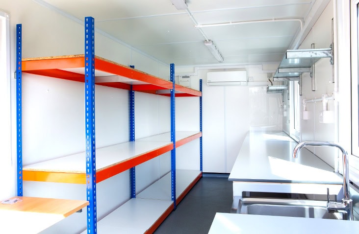 Interior of a converted shipping container fitted with long industrial storage shelves on the left and a countertop with sink and wall-mounted shelving on the right, brightly lit with white clean walls and windows.