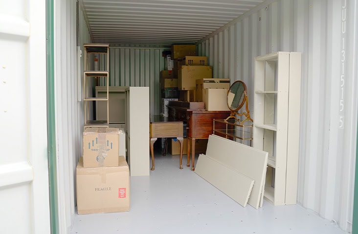 Interior of a shipping container used for storage, filled with furniture, cardboard boxes and household items.