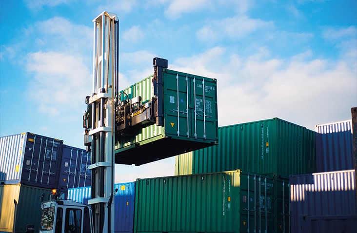 A container handler lifts a green shipping container in a storage yard, with stacks of other containers in various colours visible in the background under a bright, partly cloudy sky. 