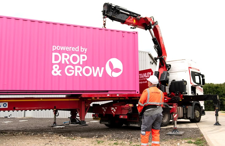 Pink shipping container with ‘Powered by Drop & Grow’ branding being lifted by a red hydraulic crane mounted on a white flatbed truck. A worker in high-visibility orange safety gear stands nearby on a gravel surface during container delivery. 