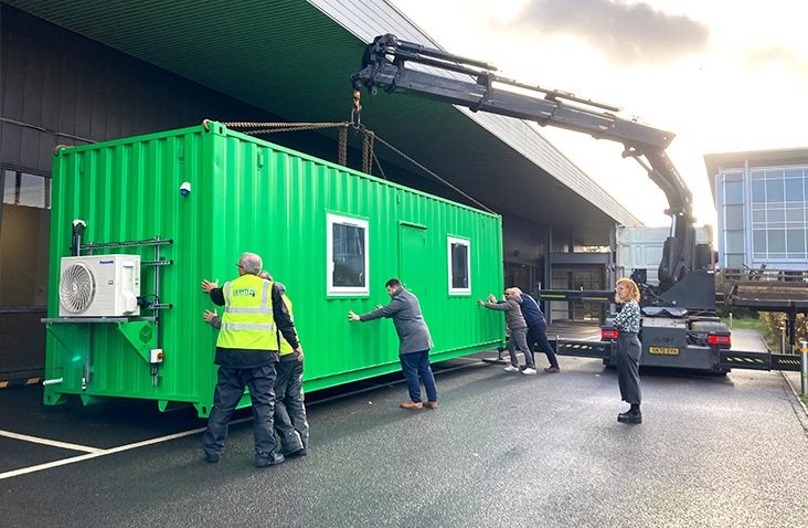 A group of workers guide a bright green modified shipping container into position as it is lifted by a crane truck outside a commercial building.
