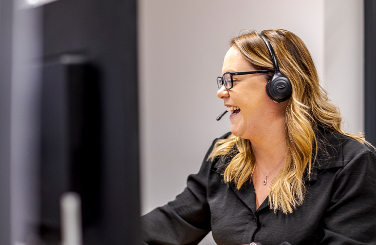 The image shows a woman wearing a headset, sitting at a desk and smiling while looking toward a computer screen. She has long, wavy blonde hair and is wearing glasses and a dark shirt. The setting appears to be an office or customer service environment, and she looks engaged and enthusiastic, suggesting positive interaction or teamwork. 