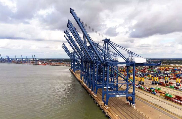 Aerial view of Felixstowe container port with tall blue cargo cranes lined along the dockside beside the sea, with stacks of colourful shipping containers in the background.