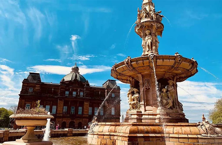 Historic Doulton Fountain in Glasgow Green with the People’s Palace museum building in the background under a bright blue sky.