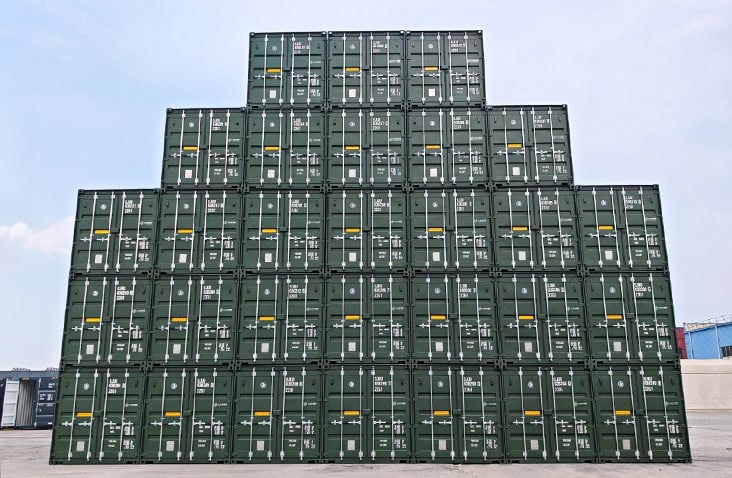 Large pyramid-shaped stack of new green S Jones shipping containers arranged in rows at a depot, ready for sale or hire.