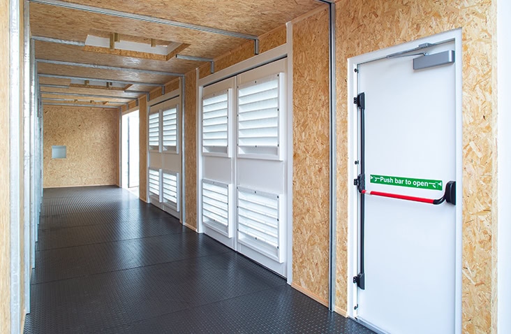 Insulated shipping container interior with OSB lining, steel anti-slip flooring, louvered ventilation panels and a fire exit door with push bar.