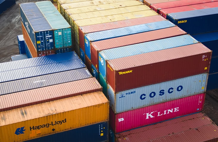  Aerial close-up view of stacked shipping containers in various colours, including blue, orange, red, and green, showing branded units such as Hapag-Lloyd, COSCO and K-Line at a busy UK freight depot.