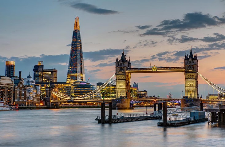 Evening view of London’s Tower Bridge and The Shard skyline reflected on the River Thames, with city lights illuminated against a dusk sky.