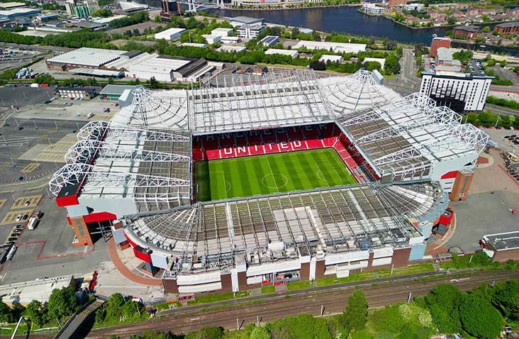 Aerial view of Old Trafford football stadium in Manchester, showing the pitch, seating, roof structure and surrounding city landscape. 