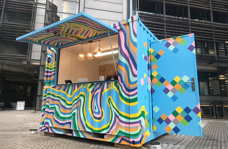 Vibrantly painted shipping container café with multicolored geometric and wavy patterns, featuring an open serving hatch and door, located in an urban outdoor setting.