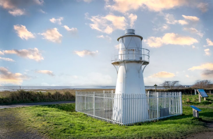 White lighthouse structure surrounded by a white metal fence on a grassy coastal area, with a cloudy sky and open landscape in the background.