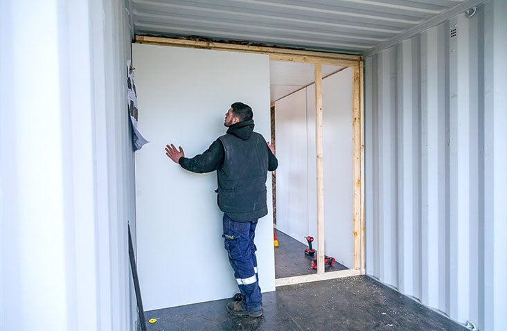 Engineer installing an internal partition wall inside a converted shipping container, fitting white insulated panelling onto a timber framework for a container modification project.