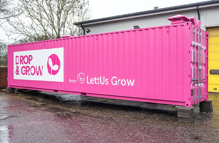Large pink 40ft shipping container with ‘Drop & Grow from LettUs Grow’ branding, positioned outdoors on concrete blocks near a building with trees in the background.