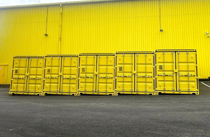 Six bright yellow shipping containers lined up side by side in front of a matching yellow industrial building, placed on a black asphalt surface.