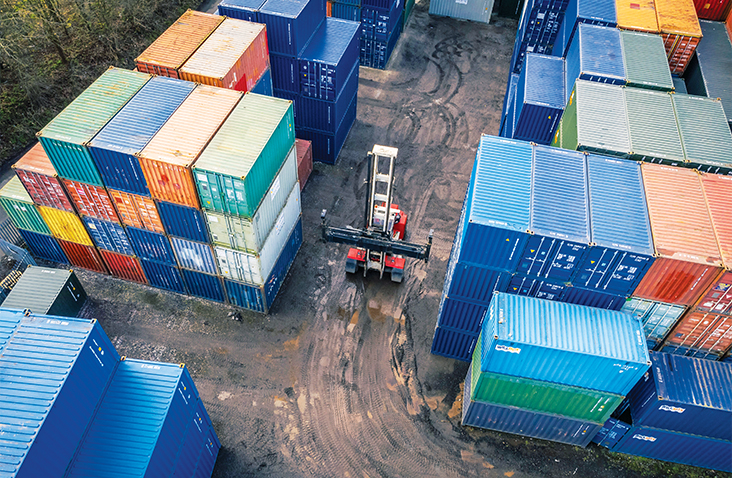Aerial view of a container storage yard with stacks of multicoloured shipping containers and a forklift truck moving between them on a dirt surface.  