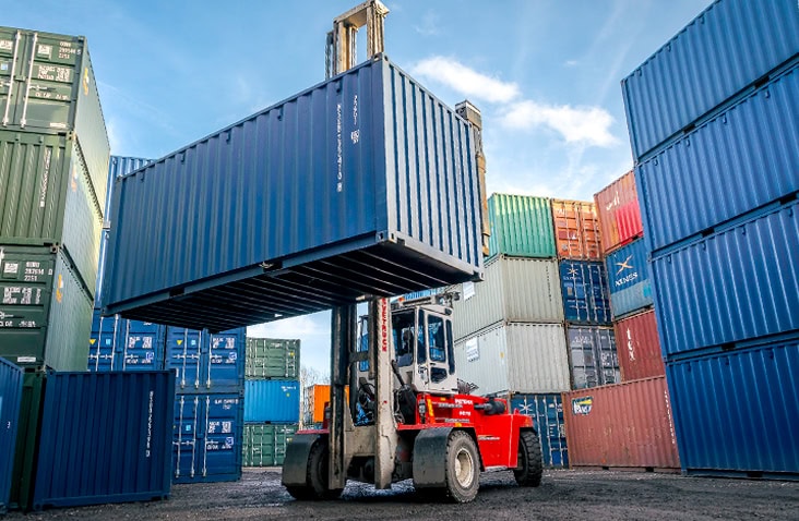 An image shows a large red forklift truck lifting a blue shipping container in an outdoor shipping container depot located in Aldridge, Walsall. The truck is surrounded by stacks of multi-coloured shipping containers. The sky is bright and clear, and the scene conveys industrial activity, logistics, and storage operations. 