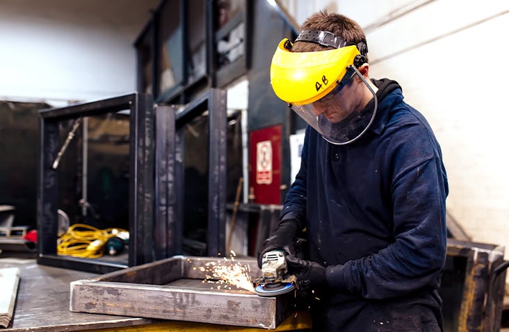 A workshop engineer wearing a yellow face shield and dark protective clothing uses an angle grinder to cut or smooth a steel frame, producing sparks on a workbench inside a metal fabrication workshop.
