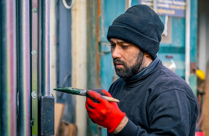 Worker wearing red gloves and a black hat carefully repainting the locking gear on a shipping container door as part of refurbishment and repair. 