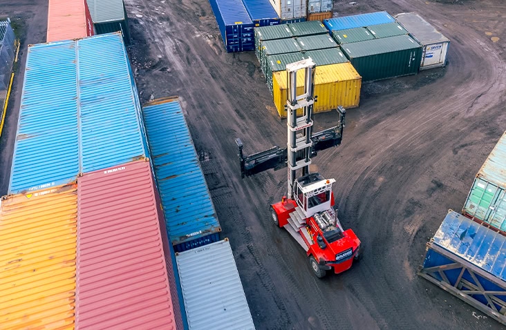 Overhead view of a red container handler forklift moving between rows of stacked shipping containers in various colours at a storage depot.