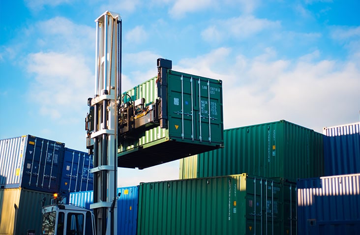 A forklift lifts a green shipping container high above stacked containers in a storage yard, under a bright blue sky