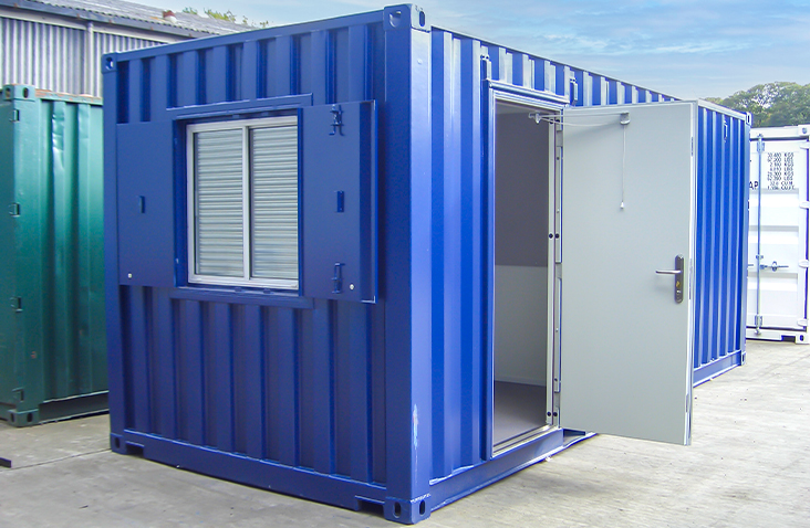 Blue 20ft converted shipping container office unit with a personnel door open and a window fitted with external steel shutters, positioned on a concrete yard.