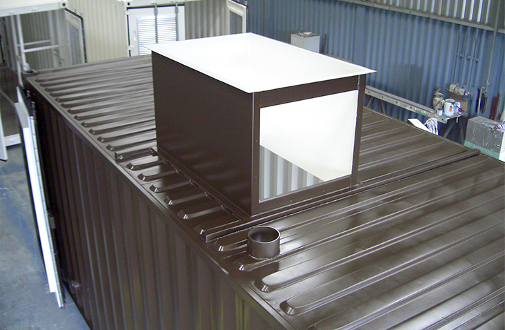 Roof of a converted shipping container fitted with a large boxed boiler flue housing. The flue enclosure is rectangular with an open top section and is positioned centrally on the container roof. The container is dark brown and located inside a workshop.