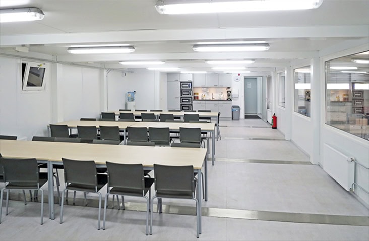 Clean modular canteen with rows of tables and chairs, a kitchenette area at the back with microwaves and coffee machines, and bright overhead lighting.
