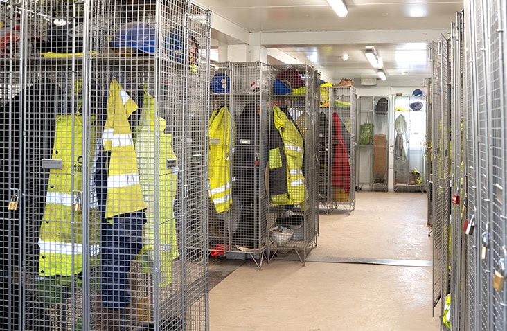 Interior of a modular changing room with metal mesh lockers holding high-visibility jackets, helmets, and workwear, brightly lit with overhead lighting.