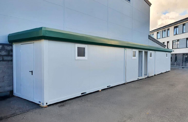 Long modular changing room building with white panelled walls, small windows, a green roof trim, and multiple doors, installed alongside an industrial facility.